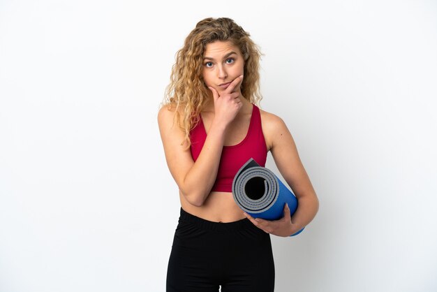 Young sport blonde woman going to yoga classes while holding a mat isolated on white background thinking