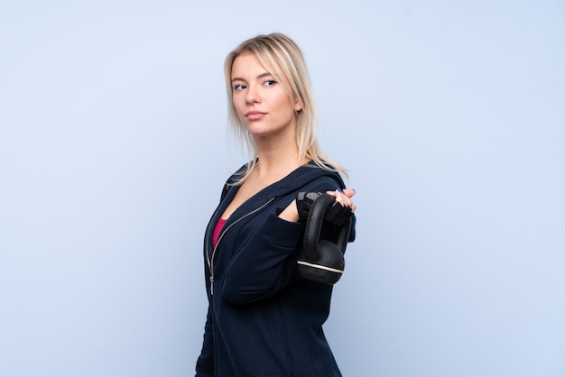 Donna bionda di giovane sport sopra la parete blu isolata che fa sollevamento pesi con kettlebell