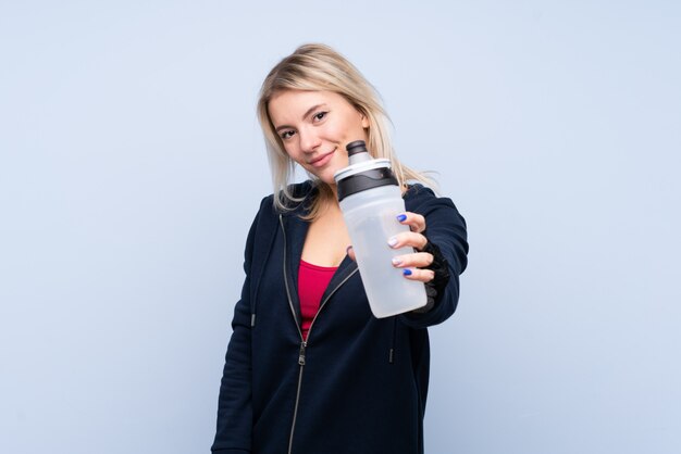 Donna bionda di giovane sport sopra la parete blu isolata con la bottiglia di acqua di sport