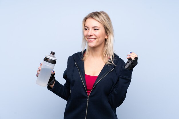Young sport blonde woman over isolated blue wall with sports water bottle