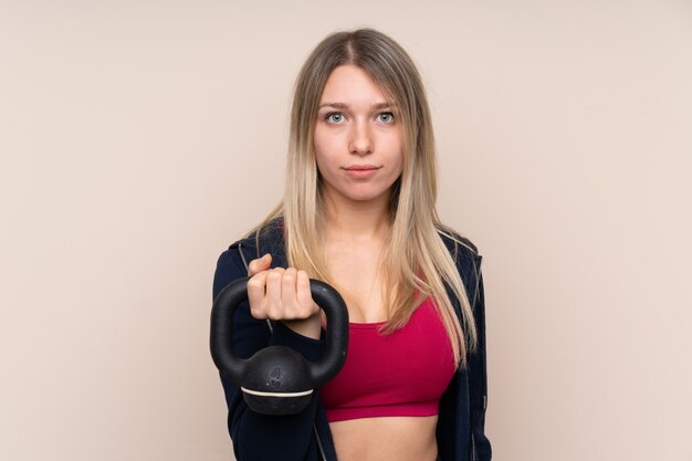 Young sport blonde woman over isolated wall making weightlifting with kettlebell and looking to the front