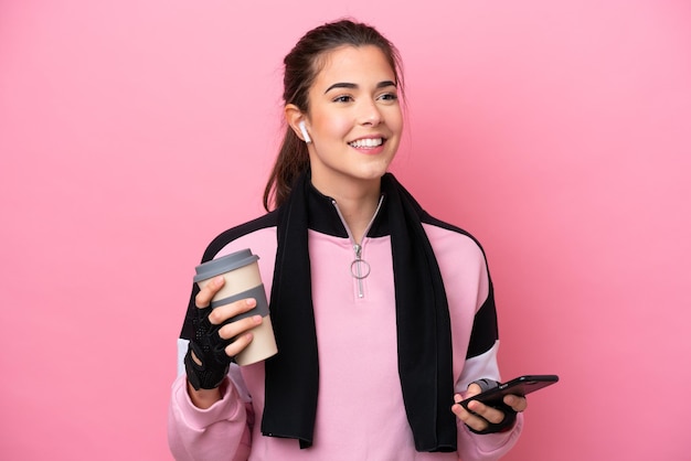 Young sport Brazilian woman isolated on pink background holding coffee to take away and a mobile