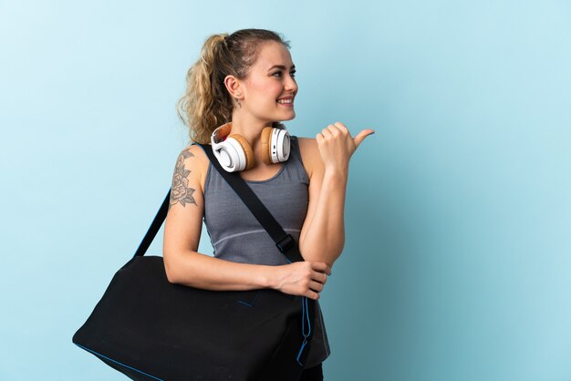 Young sport Brazilian woman with sport bag isolated on blue pointing to the side to present a product