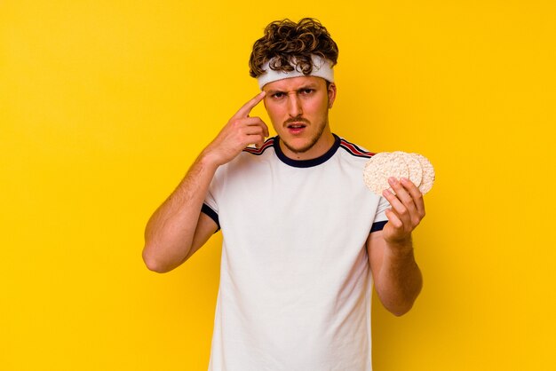 Foto uomo caucasico di giovane sport che mangia una torta di riso isolata su fondo giallo che mostra un gesto di delusione con l'indice.