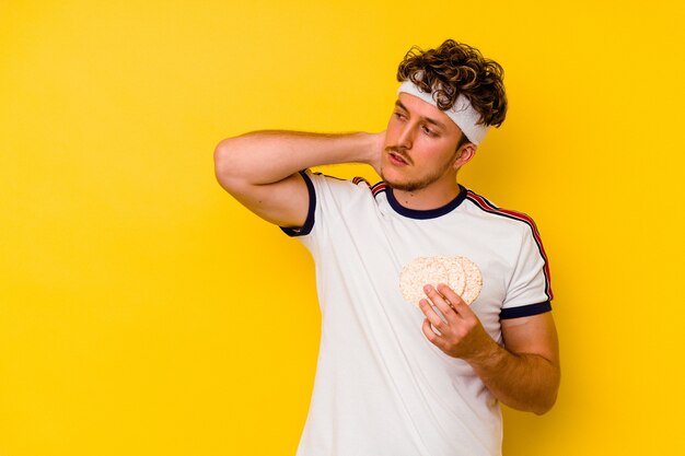 Young sport caucasian man eating a rice cake isolated on yellow background touching back of head, thinking and making a choice