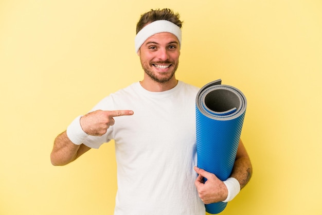 Young sport caucasian man going to yoga classes while holding a mat isolated on yellow background person pointing by hand to a shirt copy space proud and confident