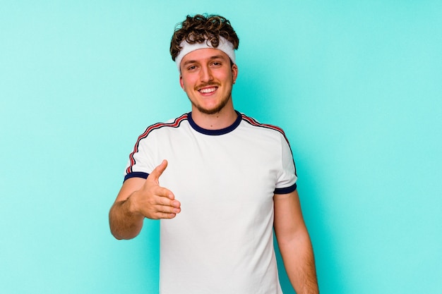 Young sport caucasian man isolated on blue background stretching hand at camera in greeting gesture.