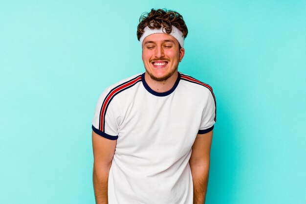 Young sport caucasian man isolated on blue wall laughs and closes eyes, feels relaxed and happy.