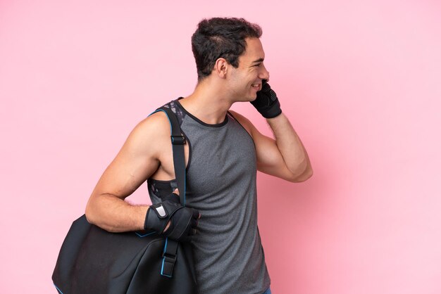 Young sport caucasian man with sport bag isolated on pink\
background keeping a conversation with the mobile phone with\
someone