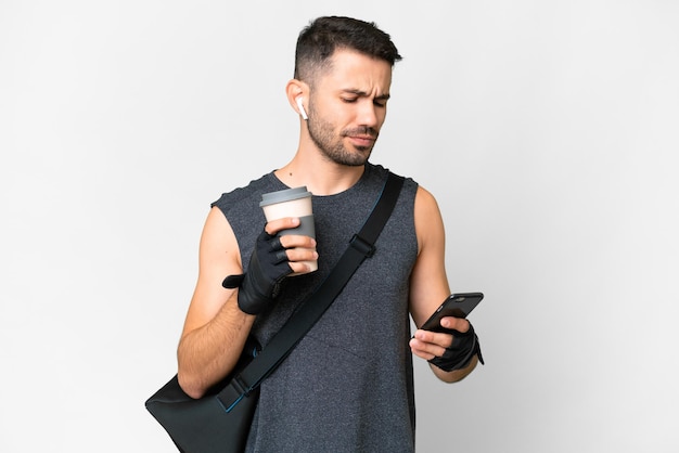 Young sport caucasian man with sport bag over over isolated white background holding coffee to take away and a mobile