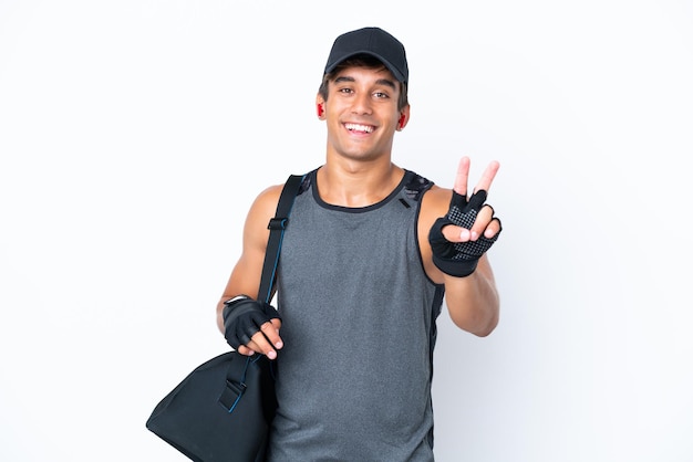 Молодой спортивный кавказец со спортивной сумкой на белом фоне улыбается и показывает знак победы