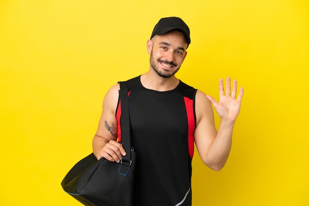 노란색 배경에 격리된 스포츠 가방을 들고 행복한 표정으로 손으로 경례하는 젊은 스포츠 백인 남자