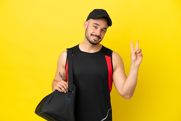 Молодой спортивный кавказец со спортивной сумкой, изолированной на желтом фоне, улыбается и показывает знак победы