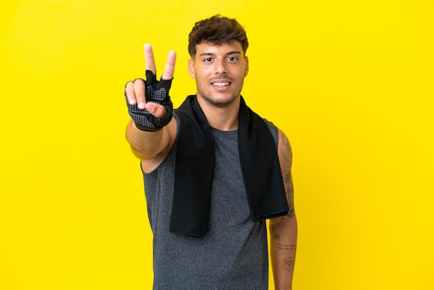 Young sport caucasian man with towel isolated on yellow background smiling and showing victory sign