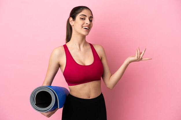 Young sport caucasian woman going to yoga classes while holding a mat extending hands to the side for inviting to come