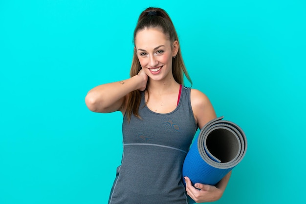 Young sport caucasian woman going to yoga classes while holding a mat isolated on blue background laughing