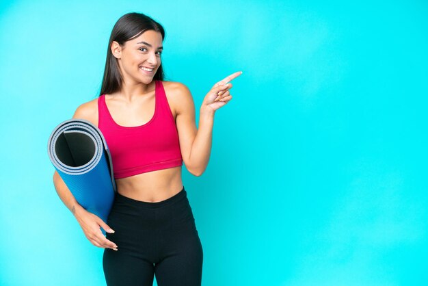 Young sport caucasian woman going to yoga classes while holding a mat isolated on blue background pointing finger to the side
