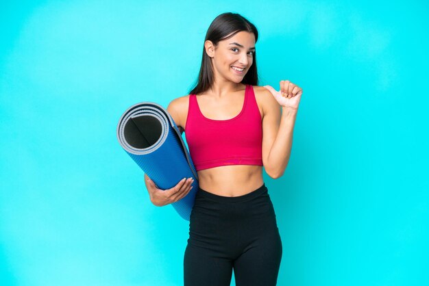 Young sport caucasian woman going to yoga classes while holding a mat isolated on blue background proud and selfsatisfied