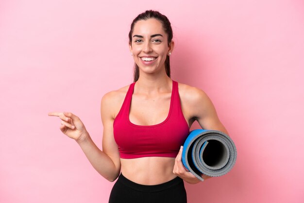 Young sport caucasian woman going to yoga classes while holding a mat isolated on pink background pointing finger to the side