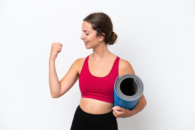 Giovane donna caucasica sportiva che va a lezioni di yoga mentre tiene un tappetino isolato su sfondo bianco che celebra una vittoria