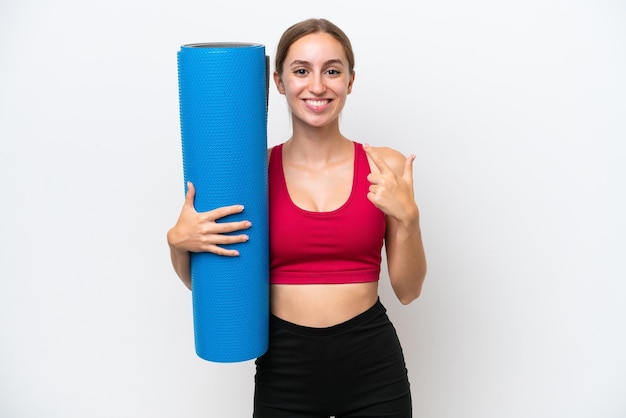 Foto giovane donna caucasica sportiva che va a lezioni di yoga mentre tiene un tappetino isolato su sfondo bianco dando un pollice in alto gesto