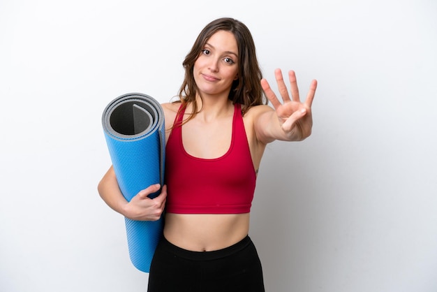 Giovane donna caucasica sportiva che va a lezioni di yoga tenendo un tappetino isolato su sfondo bianco felice e contando quattro con le dita