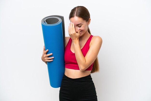 Young sport caucasian woman going to yoga classes while holding a mat isolated on white background laughing