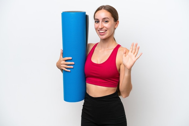 Giovane donna caucasica sportiva che va alle lezioni di yoga tenendo un tappetino isolato su sfondo bianco salutando con la mano con espressione felice