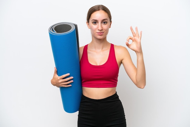 Young sport caucasian woman going to yoga classes while holding a mat isolated on white background showing ok sign with fingers