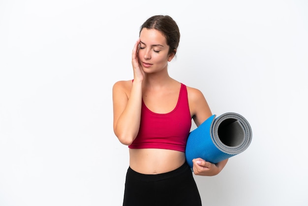 young sport caucasian woman going to yoga classes while holding a mat isolated on white background with headache