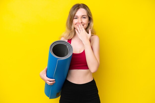 Young sport caucasian woman going to yoga classes while holding a mat isolated on yellow background happy and smiling covering mouth with hand