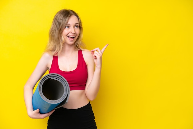 Photo young sport caucasian woman going to yoga classes while holding a mat isolated on yellow background intending to realizes the solution while lifting a finger up
