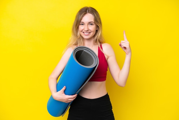 Young sport caucasian woman going to yoga classes while holding a mat isolated on yellow background showing and lifting a finger in sign of the best