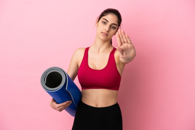 Young sport caucasian woman going to yoga classes while holding a mat making stop gesture