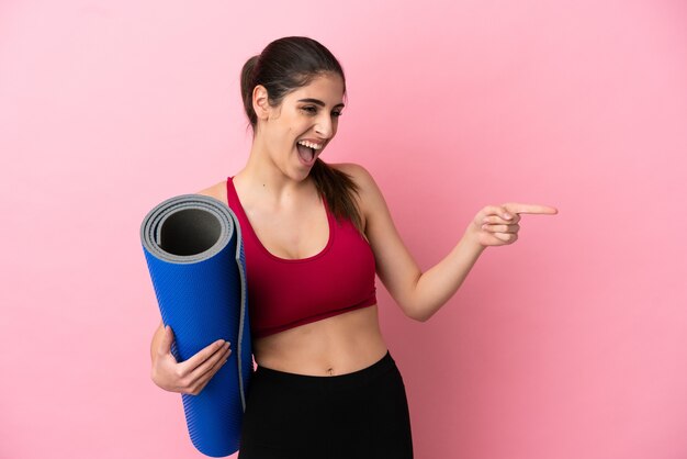 Young sport caucasian woman going to yoga classes while holding a mat pointing finger to the side and presenting a product