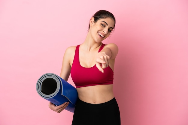 Young sport caucasian woman going to yoga classes while holding a mat pointing front with happy expression