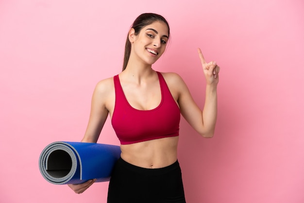 Young sport caucasian woman going to yoga classes while holding a mat showing and lifting a finger in sign of the best