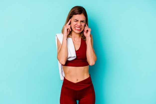 Young sport caucasian woman isolated on blue wall covering ears with hands.