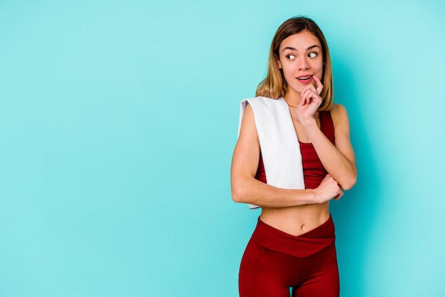 Young sport caucasian woman isolated on blue wall relaxed thinking about something looking at a copy space.