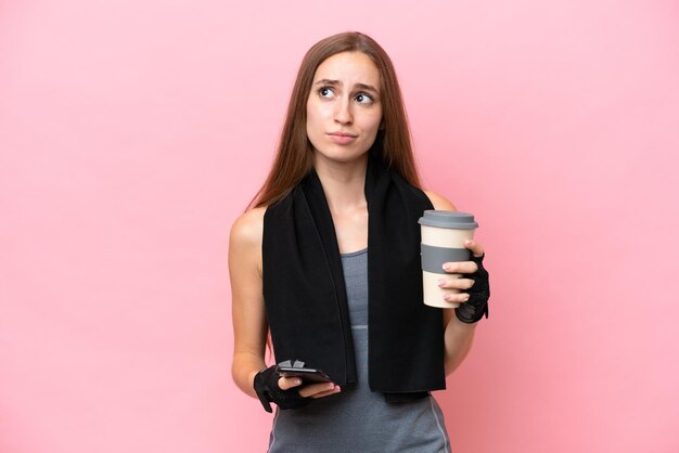 Young sport caucasian woman wearing a towel isolated on pink\
background holding coffee to take away and a mobile while thinking\
something