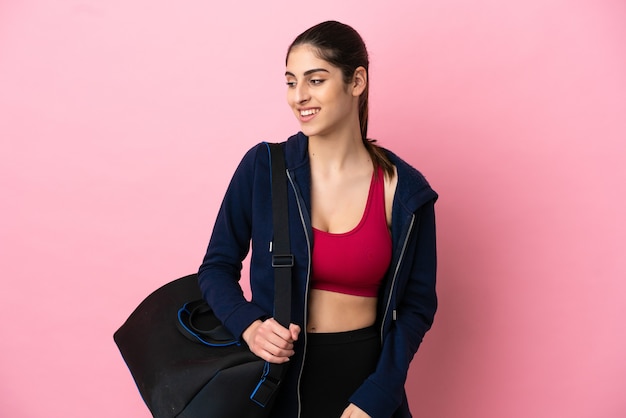 Young sport caucasian woman with sport bag isolated on pink background looking to the side and smiling