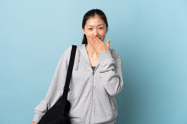 Молодая спортивная китаянка со спортивной сумкой над изолированным синим счастливым и улыбающимся прикрытием рта рукой