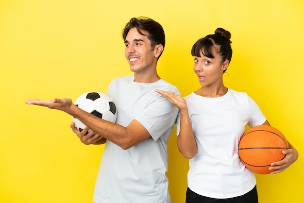 Young sport couple playing football and basketball isolated on\
yellow background extending hands to the side for inviting to\
come