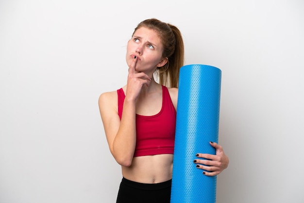 Young sport English woman going to yoga classes while holding a mat isolated on white background having doubts while looking up