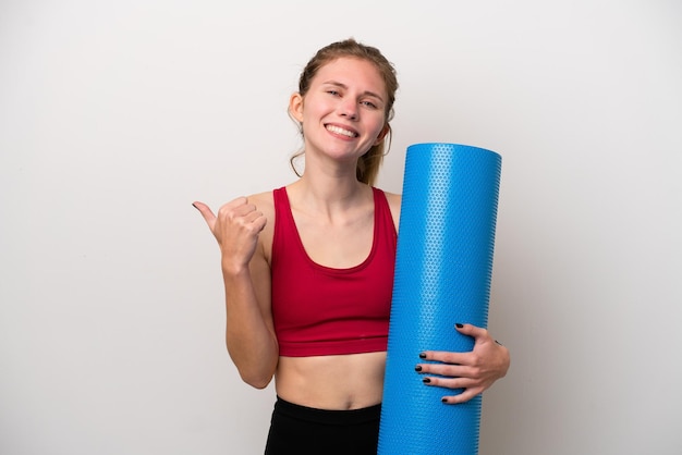 Молодая спортивная англичанка собирается на занятия йогой, держа коврик на белом фоне, указывая в сторону, чтобы представить продукт