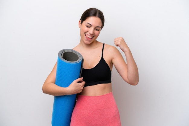 Young sport girl going to yoga classes while holding a mat isolated on white background celebrating a victory