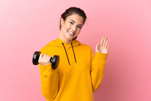 Young sport girl making weightlifting over isolated pink background saluting with hand with happy expression