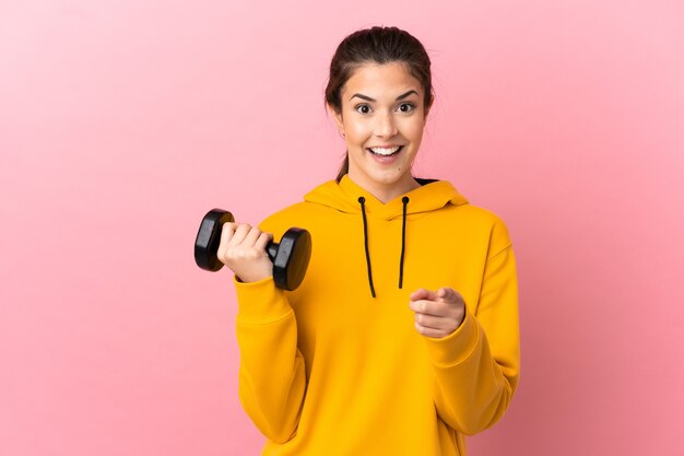 Young sport girl making weightlifting over isolated pink background surprised and pointing front