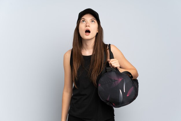Молодая спортивная девушка со спортивной сумкой изолирована на сером, глядя вверх и с удивленным выражением лица