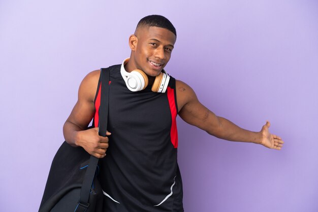 Young sport latin man with sport bag isolated  on purple extending hands to the side for inviting to come